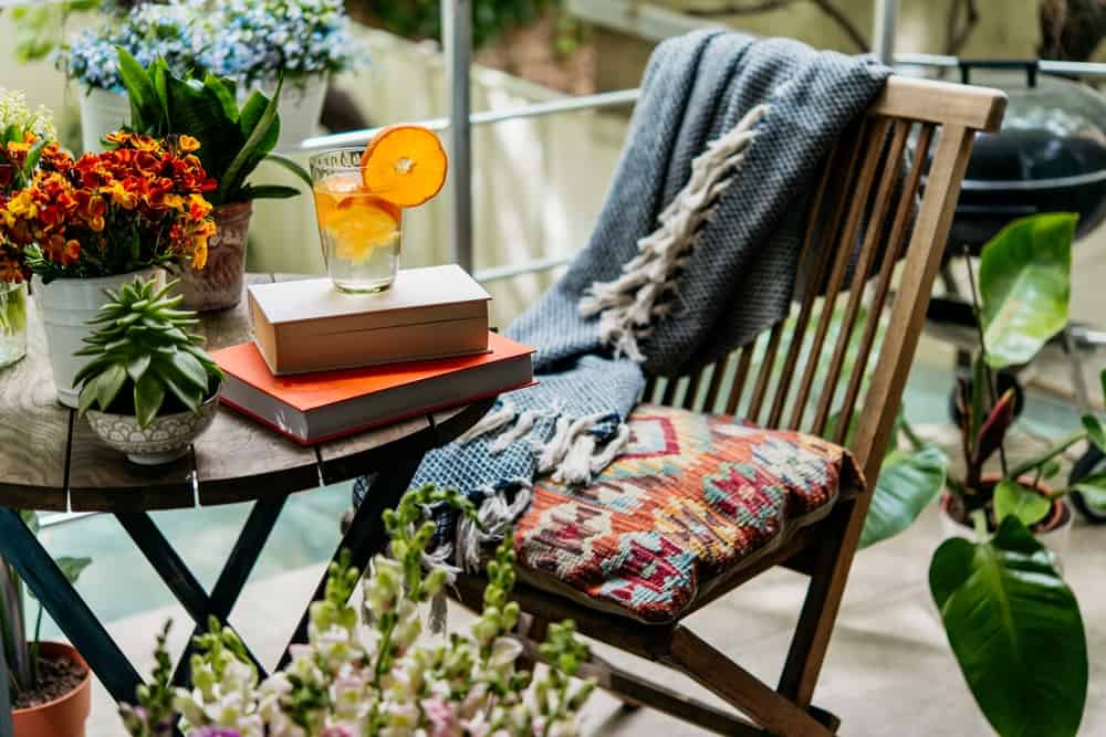A cozy patio with a chair, cushion, blanket, and table with flowers and a book and a drink