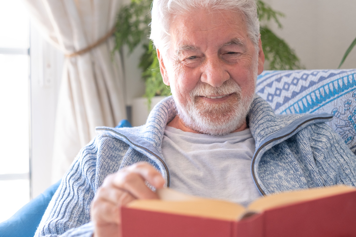 Senior 70s man seated on sofa, leisure and people concept - happy bearded senior man relaxing at home reading book